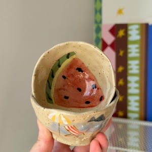 May include: Two small, beige ceramic bowls. The top bowl features a watermelon slice design. The bottom bowl has a beach scene with a beach chair and umbrella. The bowls are handmade and have a speckled texture.