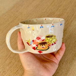 May include: A beige ceramic mug with a hand-painted design featuring a red and white checkered picnic blanket, two small cups, a lit candle, and colorful flowers. The rim is decorated with small blue and red dots, and the handle is curved.