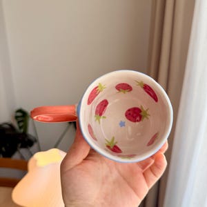 K&ouml;nnte beinhalten: Eine wei&szlig;e Keramiktasse mit blauem Rand und Henkel. Die Tasse ist mit einem Muster aus roten Erdbeeren und gr&uuml;nen Bl&auml;ttern verziert.