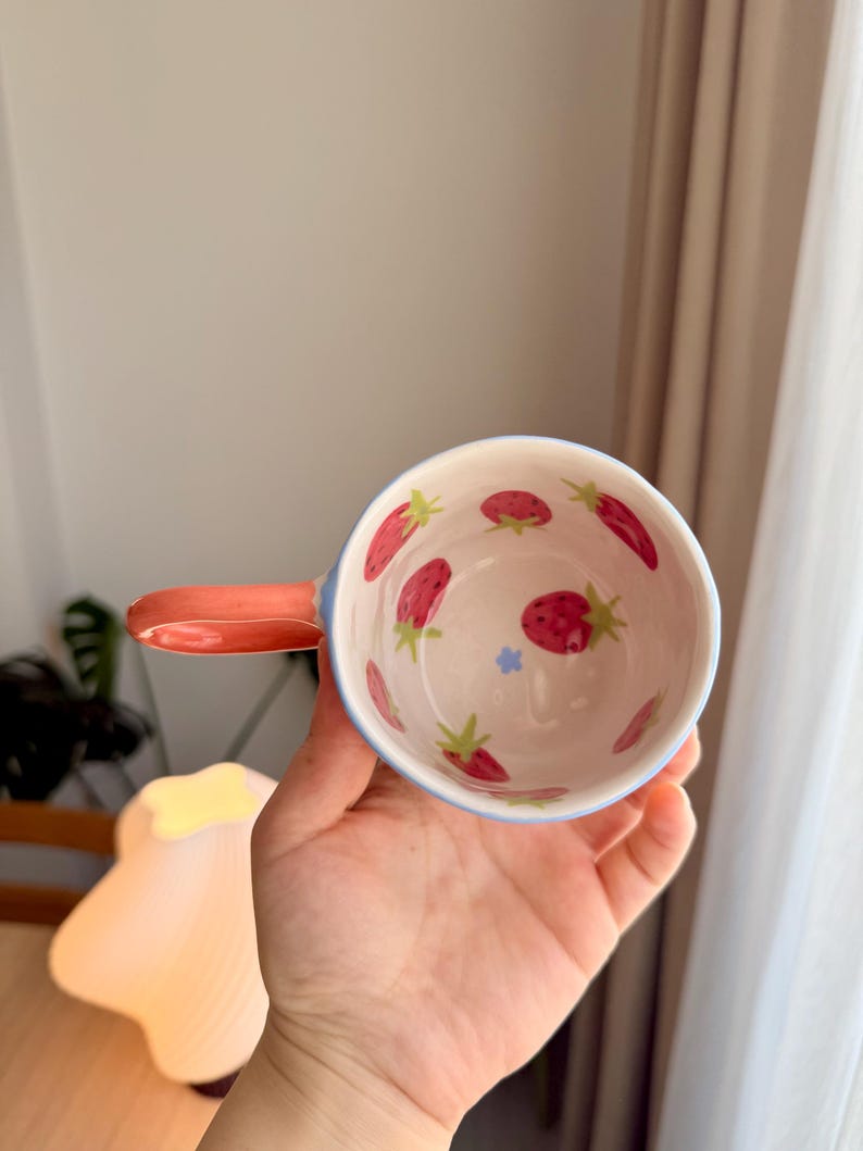 K&ouml;nnte beinhalten: Eine wei&szlig;e Keramiktasse mit blauem Rand und Henkel. Die Tasse ist mit einem Muster aus roten Erdbeeren und gr&uuml;nen Bl&auml;ttern verziert.