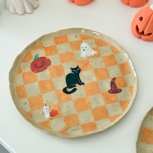Może przedstawiać: Okrągły ceramiczny talerz z nakrapianą kremową bazą i pomarańczowym wzorem w szachownicę. Talerz ozdobiony jest ręcznie malowanymi ilustracjami czarnego kota, białego ducha, czerwonej dyni, fioletowego kapelusza czarownicy oraz zapalonej świecy.