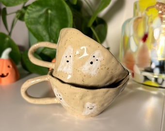 May include: Two stacked ceramic mugs with a speckled beige finish, each featuring hand-painted white ghost illustrations with small black eyes and mouths. The mugs have an irregular, organic shape with a single handle on the side of each piece.
