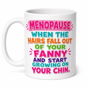 May include: White ceramic mug with the text "MENOPAUSE WHEN THE HAIRS FALL OUT OF YOUR FANNY AND START GROWING ON YOUR CHIN" in pink, blue, orange, green, and purple. The mug has a white handle.