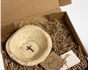 May include: A white ceramic bowl with a cross design inside, packaged in a brown cardboard box with shredded paper. The box also includes a small card with the text "Bowls Kim & Co."