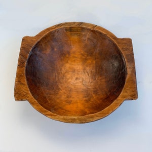 May include: A handcrafted wooden bowl with a round interior and rectangular handles. The bowl has a natural wood grain finish with varying shades of brown. The bowl is likely used for serving or display.