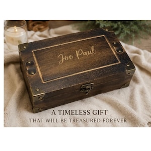 May include: A rectangular dark brown wooden box with a hinged lid, featuring brass-toned metal corner reinforcements and a central latch. The name Joe Paul is engraved in a script font on the top surface of the lid, which has a dark wood grain finish.