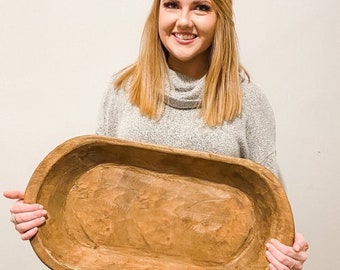 May include: A large, oval-shaped wooden dough bowl with a natural wood finish. The bowl is being held by a person.