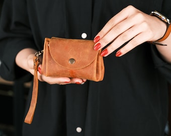 May include: A small brown leather purse with a flap closure and a snap button. The purse has a long strap and is held in a person's hand.
