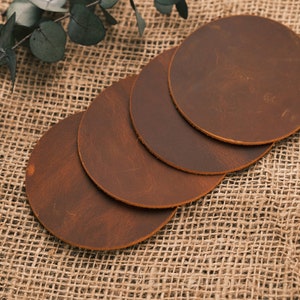 May include: Four brown leather coasters on a burlap surface. The coasters are round and have a distressed look.