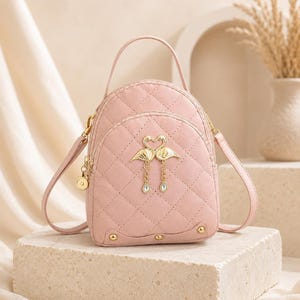 Könnte beinhalten: Ein hellrosa gesteppter Rucksack mit einem Tragegriff und verstellbaren Riemen. Die Vorderseite zeigt ein goldfarbenes Metallemblem aus zwei Flamingos, die ein Herz bilden, mit hängenden Ketten, die in kleinen klaren Kristallen enden.