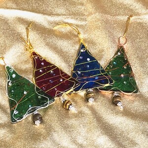 May include: Four Christmas tree ornaments in shades of green, red, and blue. Each tree is outlined in gold wire and adorned with small, sparkling embellishments. The ornaments hang from gold cords and have decorative beaded bottoms.