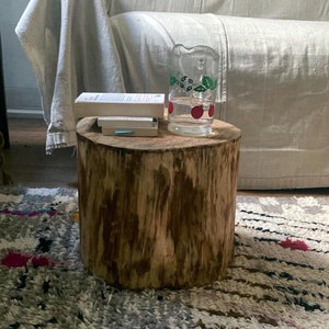 Peut inclure: Une table d'appoint en forme de souche d'arbre avec une finition en bois naturel. Un pichet en verre avec un motif floral vert et rouge est posé sur la table, ainsi qu'une pile de livres.