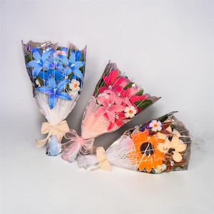 May include: Three bouquets wrapped in clear plastic and white mesh, tied with ribbons. The left bouquet features blue lilies, the center bouquet contains pink lilies, and the right bouquet displays an orange sunflower and cream-colored lilies.