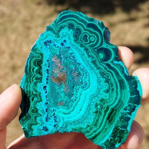 May include: A polished malachite stone with swirling patterns of deep teal, turquoise, and forest green. The natural stone has a unique, organic shape and a polished surface, showcasing its intricate mineral formations. The stone is held up against a blurred background.