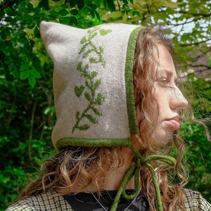 May include: A light beige knit hat with a pointed top and green trim. The hat features a green embroidered floral design on the front and green ties. The hat is a unique accessory, perfect for adding a touch of whimsy to any outfit.