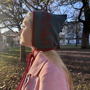 May include: A teal and red knitted hat with a pointed top and floral pattern. The hat is worn with a pink coat and red ties. This unique hat is a stylish accessory, adding a touch of colour and flair to any outfit.