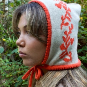 May include: A white knitted hat with a red trim and floral pattern. The hat has a long tie that can be tied under the chin.