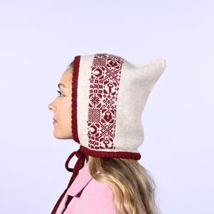 May include: A knitted winter hat with a pointed top and a burgundy trim. The hat features a cream-colored base with a repeating burgundy pattern. Burgundy ties hang from the hat. The hat is worn by a person.
