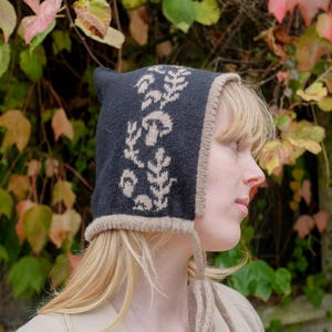 May include: A dark grey knitted hat with a beige trim and a mushroom and leaf pattern. The hat has ear flaps and ties. The background features green, red, and yellow leaves.