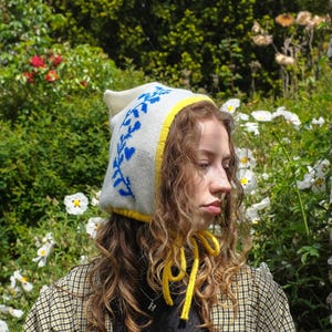 May include: A white knit hat with blue floral embroidery and a yellow trim. The hat has yellow ties and is worn with a plaid jacket. The background features green foliage and white flowers.