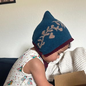 May include: A teal knitted hat with a floral design in tan and a burgundy trim. The hat is worn by a child wearing a white sleeveless top with a colorful bird pattern. The child is looking into a cardboard box.