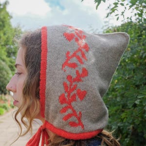 May include: A grey knitted hood featuring a red floral pattern and trim. The hood has a pointed top and ties under the chin. The design includes a repeating pattern of red leaves and hearts. The hood is made of a soft, warm material.