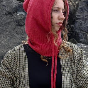 May include: A red knit hood with long ties, worn over a black top and a plaid jacket. The hood is a vibrant shade of red and appears to be made of a soft, textured knit material. The jacket has a black, white, and tan plaid pattern.