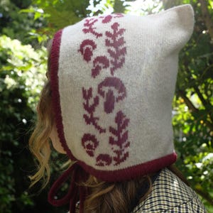 May include: A light grey knitted bonnet with a burgundy trim and ties. The bonnet features a repeating pattern of burgundy mushrooms and leaves. A unique accessory for cooler weather, this bonnet is a charming item.
