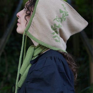 May include: A beige knitted hooded hat with green leaf embroidery and matching green scarf. The hat has a long tie that can be tied under the chin.