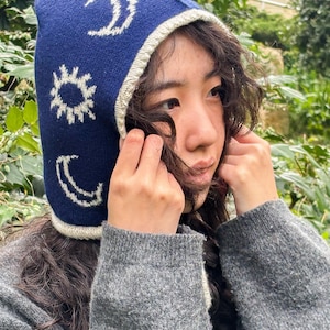 May include: A dark blue knitted hat with a pointed top and a cream-coloured trim. The hat features white embroidered celestial designs, including a sun, a crescent moon, and a full moon. The person in the image is wearing a grey sweater.