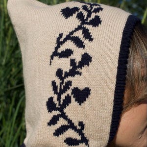 May include: A beige knitted hat with a black floral design. The hat has a pointed top and a black trim around the edge.