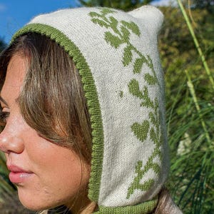 May include: A cream-colored knit bonnet with a green floral pattern and trim. The bonnet has a pointed top and ties under the chin. The background is a sunny outdoor scene with green foliage.