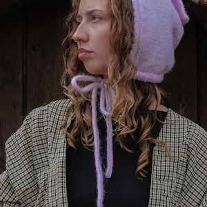May include: A light purple knit bonnet with ear flaps and long ties. The bonnet is made of a soft, fuzzy material. The person is wearing a black top and a plaid jacket.