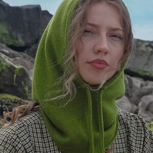 May include: A green knit hood with drawstrings. The hood is a vibrant shade of green and is worn over a plaid shirt. The background features rocks and a blue sky.