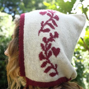 May include: A cream-colored knitted hood with a burgundy trim and floral design. The hood features a pointed top and ties, with a burgundy floral pattern on the front. The trim is a darker shade of burgundy.