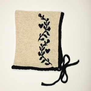 May include: A beige knit bonnet with a black floral design on the front. The bonnet has a black border and black ties. The design features a vine with heart-shaped leaves.