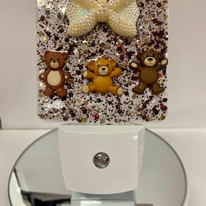 May include: A white night light with a decorative top featuring a glittery background, a cream-colored bow, and three teddy bear embellishments. The night light is designed to plug into a standard wall outlet.