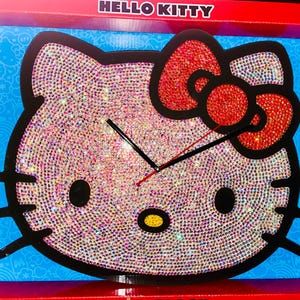 May include: A Hello Kitty clock with a black outline and a sparkling, iridescent surface. The bow is red and also covered in sparkling crystals. The clock hands are black. The clock is in a red and blue box with the words "HELLO KITTY" at the top.