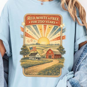 Op de afbeelding: Lichtblauw T-shirt met een afbeelding van een landelijk landschap met een boerderij, schuur en Amerikaanse vlag. De tekst "RED, WHITE & FREE &bull; FOR 250 YEARS" staat in een rechthoekige lijst. Het shirt heeft korte mouwen en een losse pasvorm.