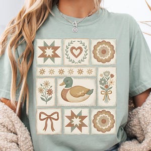 May include: A light green t-shirt featuring a patchwork design with various floral and geometric patterns. The central image is a duck. The shirt has a relaxed fit and short sleeves.