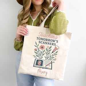 May include: Beige canvas tote bag with the text "Teaching Tomorrow's Scanners" and the name "Mary". The design includes flowers, leaves, a book with a heart, and a pencil. The bag has a light beige handle.