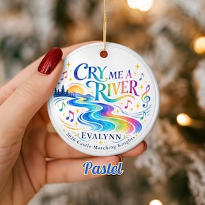 May include: A round white ceramic ornament featuring a colorful graphic of a winding river, a sun, and musical notes. The text reads "CRY ME A RIVER", "EVALYNN", and "2026 Castle Marching Knights". It hangs from a gold string against a blurred background.