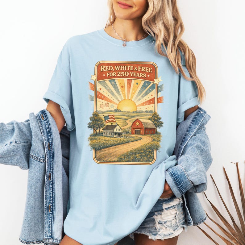 Op de afbeelding: Lichtblauw T-shirt met een afbeelding van een landelijk landschap met een boerderij, schuur en Amerikaanse vlag. De tekst "RED, WHITE & FREE &bull; FOR 250 YEARS" staat in een rechthoekige lijst. Het shirt heeft korte mouwen en een losse pasvorm.