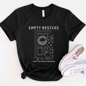 May include: Black t-shirt with the text "EMPTY NESTERS" above a door graphic with a wreath, bird, and dog. The text "JUST YOU, ME & THE DOGS" is below the graphic. White sneakers with red accents are next to the shirt.