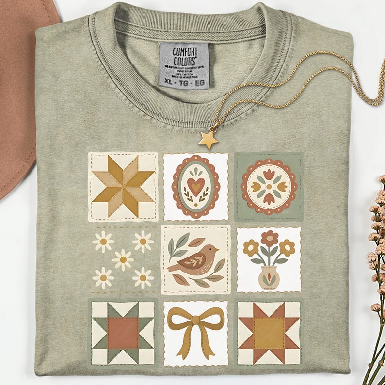 May include: A sage green t-shirt with a patchwork design featuring various floral and geometric patterns. A gold necklace with a star pendant rests on the shirt. The tag reads "Comfort Colors" and "XL - TG - EG".
