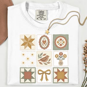 May include: White t-shirt with a patchwork quilt design featuring various floral and geometric patterns in muted colors. A gold star pendant necklace rests on the shirt. The shirt has a Comfort Colors tag, size XL-TG-EG.