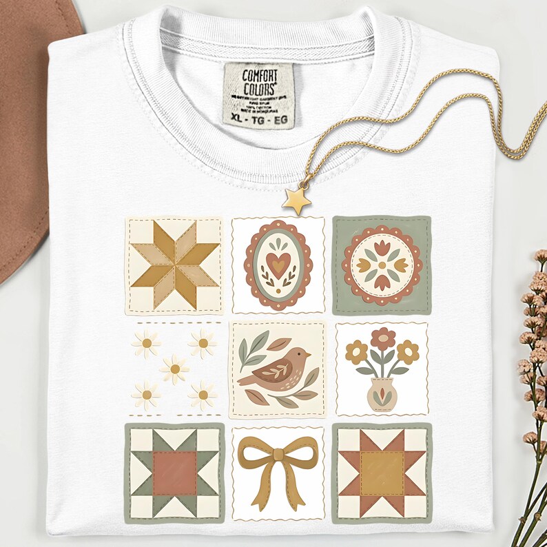 May include: White t-shirt with a patchwork quilt design featuring various floral and geometric patterns in muted colors. A gold star pendant necklace rests on the shirt. The shirt has a Comfort Colors tag, size XL-TG-EG.