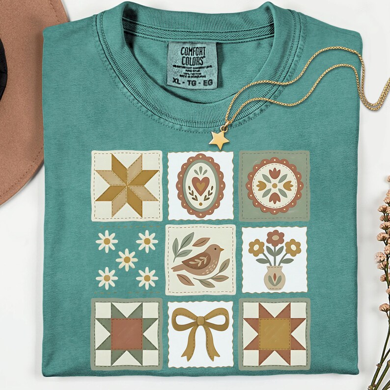 May include: Teal-colored t-shirt with a patchwork design featuring various floral and geometric patterns in muted colors. A gold necklace with a star pendant rests on the shirt. The shirt has a Comfort Colors tag, size XL-TG-EG.