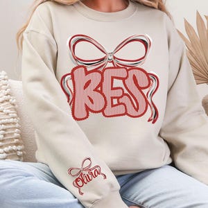May include: Beige sweatshirt with a red and white striped "BES" design, topped with a bow. The sleeve features a bow and the name "Olivia" in red script. The sweatshirt is a casual, comfortable style.