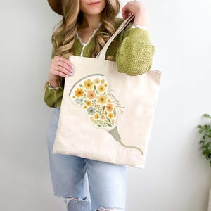 May include: Beige canvas tote bag featuring a floral design in yellow, orange, and green. The design is shaped like a medical ultrasound probe, with the word "Sonographer" written in script. The bag has a simple, minimalist design.